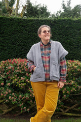 Woman in plaid top and yellow pants stands in front of floral and greenery hedges. She wears a knit grey cardigan vest with two front pockets.  thumbnail