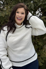 Woman in a white crochet sweater with a turtleneck and slightly puffy sleeves. The sweater has black outlines along the raglan, on the outside of the sleeves, and above the ribbing at hem and cuff.  thumbnail