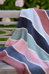 Striped knit blanket draped over a park bench. The blanket has stripes of brick red, blue, pale pink, sage green, navy, and sky blue separated by a narrow stripe of cream.  thumbnail