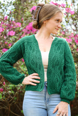 Woman stands in front of a flowering shrub, wearing an emerald green cabled crochet cardigan, cream tank top, and blue jeans.  thumbnail