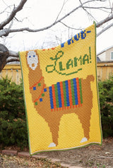 Afghan draped over tree branch in a backyard. The afghan has a yellow background with a tan llama, with a blue-orange-green and striped blanket on its back. The phrase "No Prob-Llama" is written above the llama's back.  thumbnail