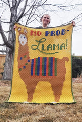 Woman holds afghan in front of her, toward camera. The full llama can be seen on the front, including its pale face and inner ears.  thumbnail