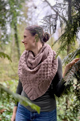 Woman stands in a lush pine forest, wearing a tan cabled shawl bandana style.  thumbnail