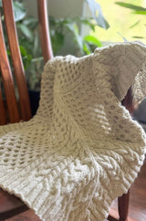 Cream cabled knit blanker draped over wooden chair.  thumbnail