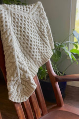 Cabled knit blanket knit in cream from the center out. Blanket is draped over the back of a wooden chair.  thumbnail