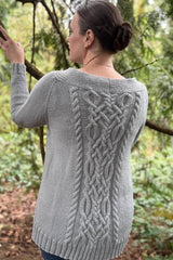 Back view of woman standing in conifer forest. She wears a pale grey cardigan with a large cabled panel down the back.  thumbnail