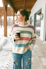 Woman stands on a covered porch with a snowy background. She wears a striped sweater, casual jeans, and holds a coffee cup. The sweater is knit in cream with stripes of dusty purple, sage green, peachy pink, and sky blue.  thumbnail