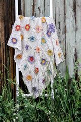 Crochet blanket on a white wooden ladder, propped against a weathered wood building with long grass. The blanket is white with multicolored flower embellishments.  thumbnail
