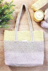 Flat lay of crochet tote bag, on a wooden table top with greenery to the side as well as a candle and skeins of yarn. The bag has a woven texture and large stripes of pale yellow, beige, and palest purple.  thumbnail