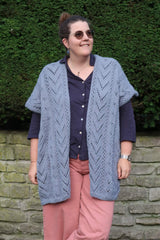 Woman wears a light blue lace cardigan vest over a dark blue top and coral pants. She stands in front of a stone wall topped by a hedge.  thumbnail