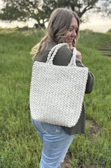 Woman stands in a green field with a crochet tote bag slung over her shoulder. The bag is a pale green and has a textured stitch that looks woven  or braided.  thumbnail