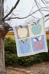 Crochet afghan draped over a tree branch in a backyard. The afghan had 4 apples, each in a different color on a contrasting background.  thumbnail