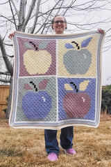 Woman holds square crochet afghan, with 4 apples, one each in pale yellow, sage green, blue, and dusty red. Each apple is on a contrasting background. There is a striped border around the entire afghan.  thumbnail