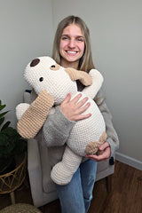 Young woman holds stuffed dog. The dog toy/pillow is as big as her torso, with a white body and light brown ears and a patch around the tail. She stands indoors, in front of a grey armchair.  thumbnail