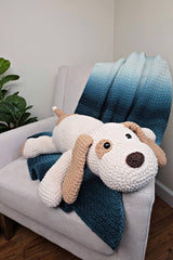 Pillow sized crochet stuffed dog resting on a blue blanket on an armchair. The dog resembles a beagle, with a white body, tan ears, tail, and a patch around one eye.  thumbnail
