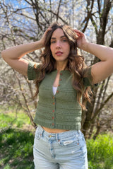 Woman stands on trail with grass and saplings in the background. She wears denim shorts and a green cardigan, buttoned partway up.  thumbnail