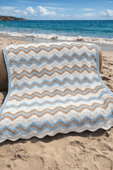 Crochet ripple blanket in stripes of white sky blue, and warm tan. It's photographed on a plush couch, and a generative AI background has placed it on a sunny beach.  thumbnail