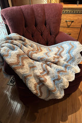 Crochet ripple blanket is laid over a smaller armchair, on a wooden floor.  thumbnail