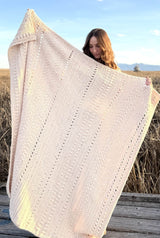 Woman standing on boardwalk in a wetland area. She holds up a crocheted throw in a cream colored yarn, with bands of different textures.  thumbnail