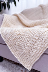Close up of knit cream colored blanket laying on grey couch. The blanket is made in garter stitch and seed stitch.  thumbnail