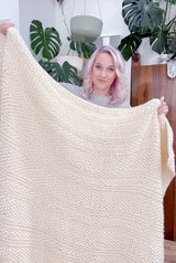 Woman with pale pink hair holds up a cream colored throw blanket, standing in front of several monstera plants. The blanket has stripes of seed stitch and garter stitch texture.  thumbnail