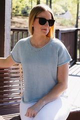 Woman sitting on outside deck, in sunglasses and white jeans. She wears a pale blue knit t-shirt with a ribbed collar and rolled cuffs on short sleeves and hem.  thumbnail