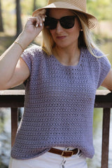 Woman stands on an outdoor deck, wearing a hat, sunglasses, and white jeans. She wears a crochet sleeveless top in a pale blue-purple.  thumbnail