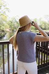 Side back view of sleeveless crochet top in a textured stitch pattern. The top is a pale blue-grey-purple. She wears a bucket style sun hat and white jeans, standing on a deck overlooking water.  thumbnail