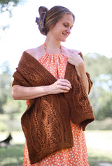 Woman in a floral summer dress wears a crochet cabled wrap in a warm caramel brown.  thumbnail