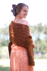 Back view of rectangular crochet wrap, with a diamond cable pattern. The wrap is a caramel brown color, worn over a summer orange and white floral dress by a woman standing outdoors.  thumbnail