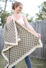 Woman standing in backyard in front of wooden fence, wearing a cream top and jeans. She holds a crochet throw blanket in a colorwork pattern. The blanket is made of cream and olive green.  thumbnail