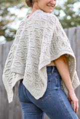 Back view of cabled crochet poncho The poncho is made in a cream color and is worn by a  woman standing in a backyard in front of a wooden fence.  thumbnail