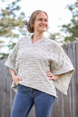 Front view of crochet poncho, made in a cream color yarn and worked side to side in a textured cable pattern. It's worn by a woman in jeans, standing in a backyard with trees and a fence in the background.  thumbnail