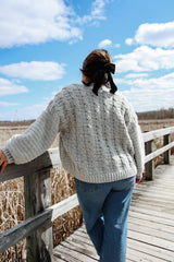Back view of crochet cardigan. It has cables on the back and plain sleeves. It is worn by a woman in jeans, with a bow in her hair, standing on a boardwalk above a wetland.  thumbnail