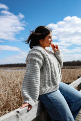 Side view of crochet cardigan, showing cables on front and plain sleeves. The woman wearing it is in jeans, siting on the railing of a boardwalk above a wetland area.  thumbnail
