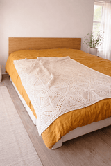 Cream crochet blanket draped across mustard orange coverlet on bed. The walls in the room are white and there is a plant in front of a window behind the bed.  thumbnail
