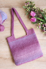 Crocheted tote bag in shades of lilac and purple laid on a pale wooden surface. Two skeins of yarn peek from the left side, and a mixed floral bouquet is on the right.  thumbnail