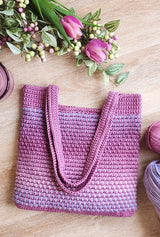 Crocheted tote bag with shoulder straps folded over the front of the bag. A mixed bouquet is at the top of the photo, and both bag and flowers are on a pale wooden surface.  thumbnail