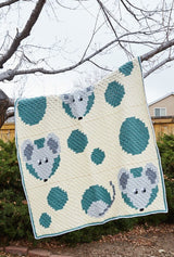 Crocht afghan hangs over tree branch in late-spring backyard. The afghan is worked in blocks and has threey grey mice face peeking through grey-green holes of "cheese" on a cream background.  thumbnail