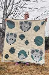 Woman holds crochet afghan out toward camera, standing in a spring backyard. The blanket is worked in shades of cream, grey-green, and grey.  thumbnail