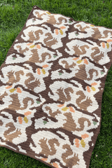 Crochet blanket laid out on grass. The blanket is made in cream and brown with pops of orange, green, and yellow. It features scenes of squirrels in varied poses.  thumbnail
