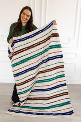 Woman holds a crochet afghan out to the side, standing in a bare room. The afghan is made in cream with stripes of green, blue, and brown.  thumbnail
