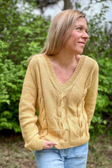 Woman standing in front of a greenery hedge wears a butter yellow sweater and blue jeans. The sweater is knit in columns of stockinette with three wide cables on the front, and has a v-neck with a ribbed edge.  thumbnail