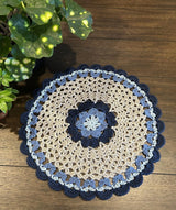 Crochet doily laid on a wooden table, with a spotted green plant nearby. The doily is made in light, medium, and dark blue, and cream.  thumbnail