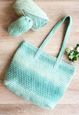 Pale green crochet tote bag laid flat on a pale wooden surface. A sprig of pale yellow flowers is laid to the right, and the yarn balls are in the upper left.  thumbnail