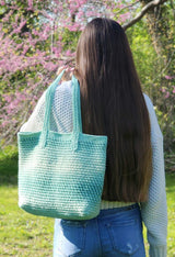 Woman with long dark hair, a cream sweater, and jeans stands outdoors. She holds a pale green crochet tote over her left shoulder.  thumbnail