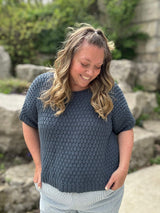 Woman standing in front of boulders and a tree. She wears a textured knit t-shirt in a denim blue.  thumbnail