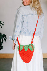 Woman in a pale denim jacket and a white dress stands indoors. She wears a crocheted purse in the shape of a strawberry, with a red berry and strap, and green leaves around the top opening.  thumbnail