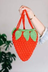 Woman holds strawberry bag toward camera, hanging by strap. The ted body has a puff stitch texture, and there are green leaves around the top.  thumbnail