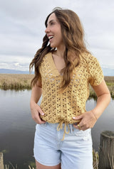 Woman in a straw yellow crochet top and pale denim shorts stands in front of a pond, with reed grasses in the background.  thumbnail